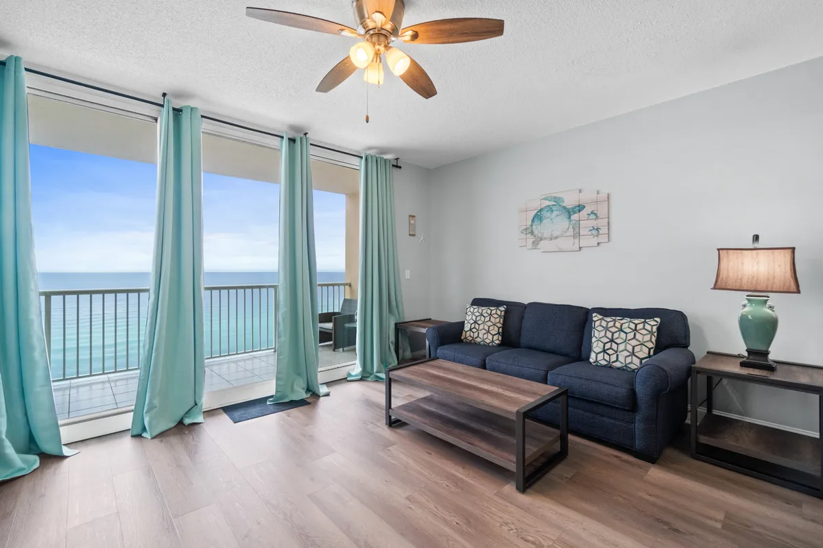 A tranquil living room flooded with natural light from expansive floor-to-ceiling windows. The blue sofa with patterned cushions, wooden coffee table, and turquoise curtains create a serene coastal ambiance. Through the window, a private balcony and sweeping ocean views are visible, emphasizing the connection between indoor comfort and the stunning beachfront setting.