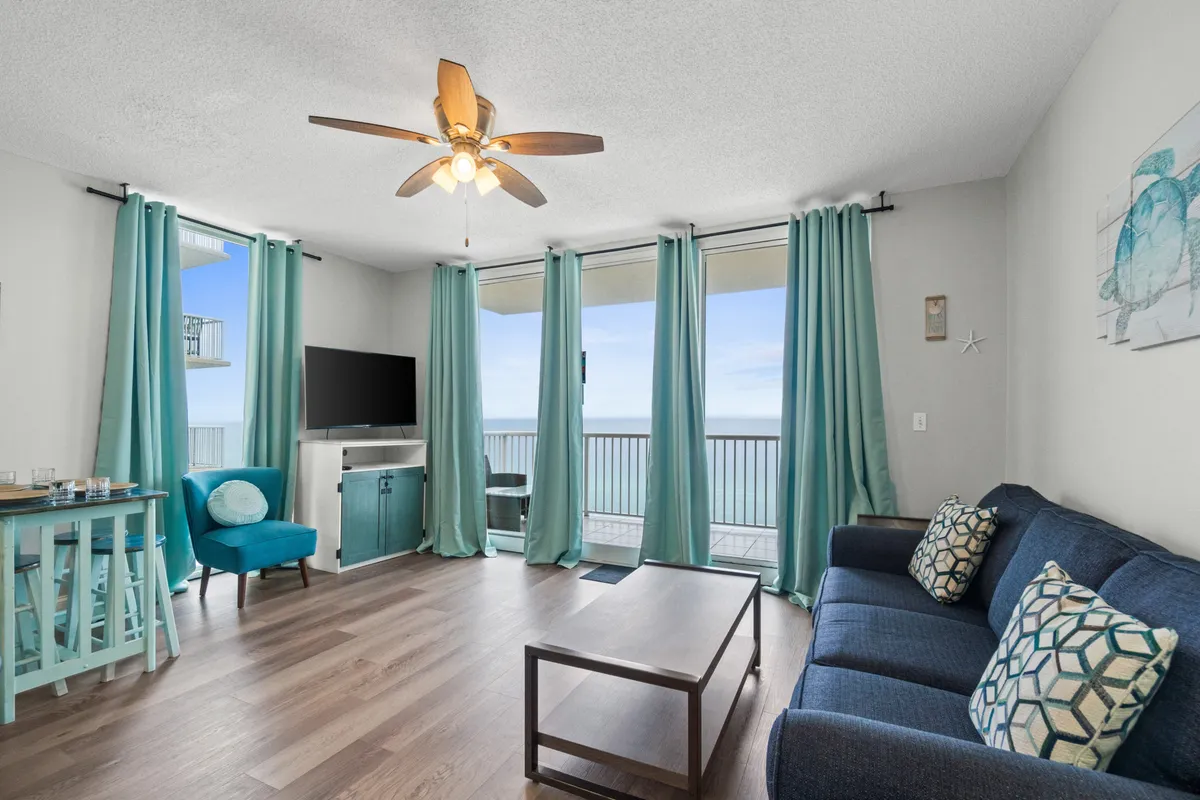 A bright, airy living area with floor-to-ceiling windows framing vibrant ocean views. The blue upholstered sofa, turquoise curtains, and matching accent chair establish a relaxed, upscale vibe. A small dining table with bar stools is set for casual meals or drinks, and the balcony is just steps away, inviting guests to transition seamlessly from inside to out.