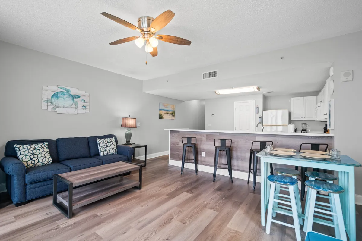 This inviting interior features a navy blue couch with patterned pillows, a kitchen island with three bar stools, and a small turquoise dining table with stools. The ceiling fan and light fixture add to the cozy atmosphere, while the overall space feels open, comfortable, and ideal for both casual dining and relaxing.