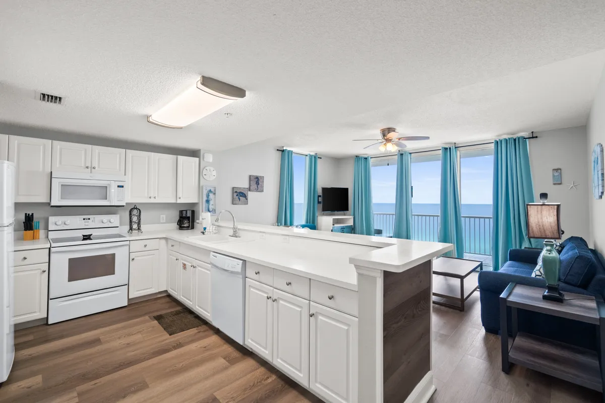 A spacious, open kitchen with white cabinetry and modern appliances occupies the left side of the image. The kitchen flows seamlessly into a cozy living space with a navy blue sofa and flat screen TV. Large sliding glass doors with turquoise curtains lead to a balcony with an ocean view, filling the area with natural light. A long, white countertop island separates the kitchen from the living area, inviting gathering and casual dining.