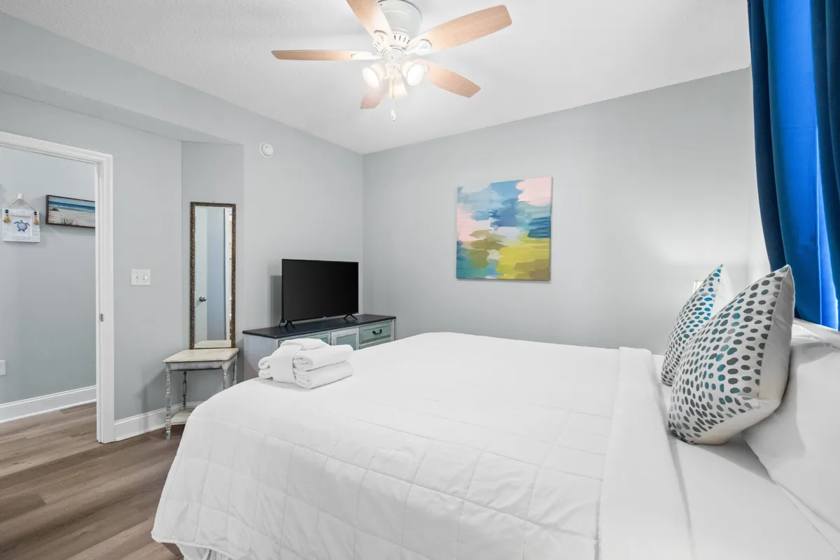 A peaceful bedroom scene with a king-size bed adorned with white linens and patterned pillows. A flat-screen TV sits atop a dresser, an abstract painting adds color, and a full-length mirror with a vintage chair provides a touch of elegance and utility.