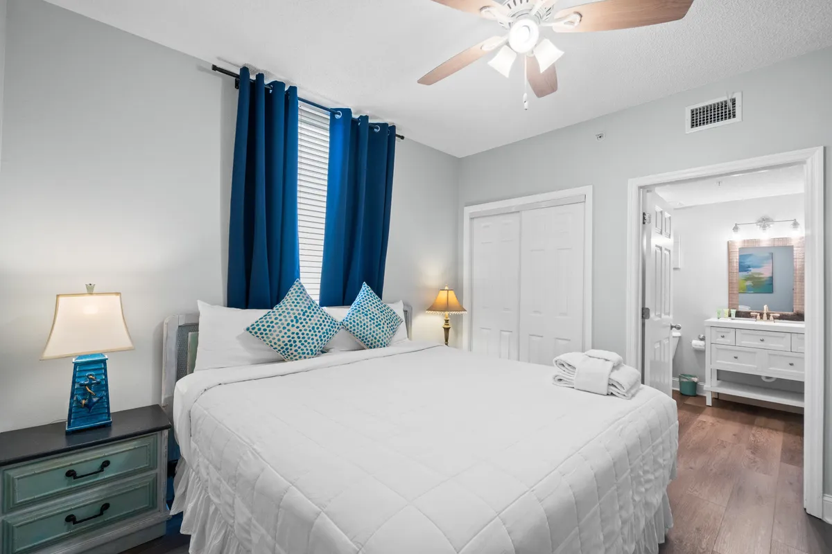 A welcoming master bedroom with a king-size bed dressed in crisp white bedding and decorative pillows. Blue curtains frame a window, flanked by nightstands with elegant lamps. The view into the adjacent bathroom is visible, showing a vanity with a mirror—illustrating the seamless connection between bedroom and en-suite.