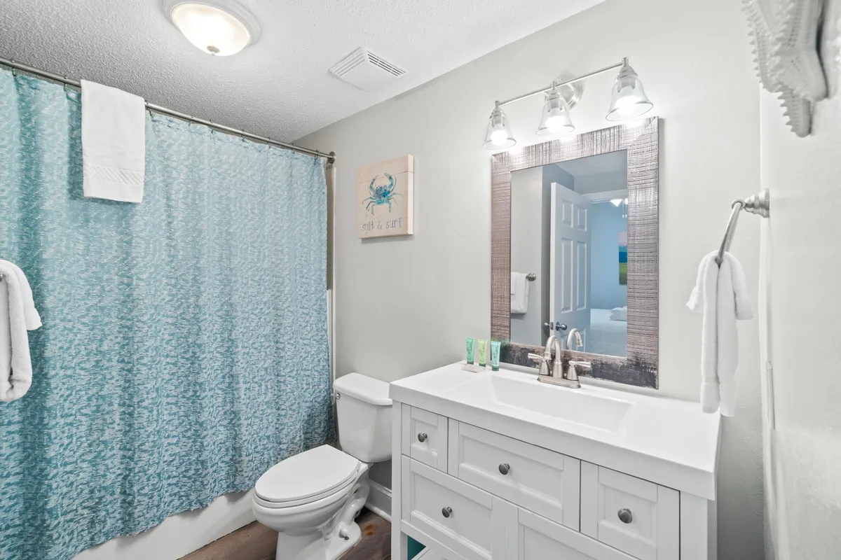 A calming bathroom with a vanity and mirror, toilet, and shower with an ocean-themed curtain. Towels are neatly arranged, creating a fresh and inviting space ideal for relaxation and refreshing routines after a day at the beach.