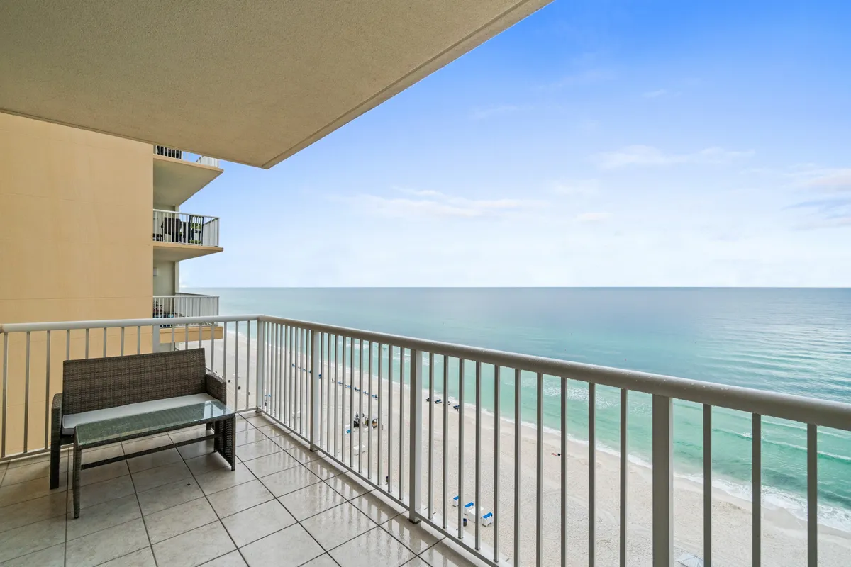 A private balcony furnished with seating overlooks an expansive ocean view. Below, the pristine white sandy beach stretches out, meeting the clear blue sky at the horizon. The scene is tranquil and inviting, perfect for quiet moments or sharing a sunset cocktail.