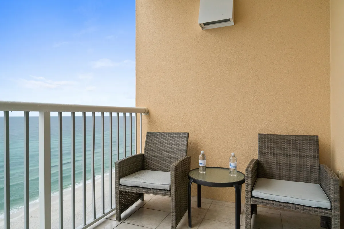 Two cushioned wicker chairs and a small round glass-top table are positioned on a balcony overlooking the ocean. The railing frames a serene view of the water, inviting guests to relax, have morning coffee, or enjoy a quiet break while taking in the Gulf breeze.