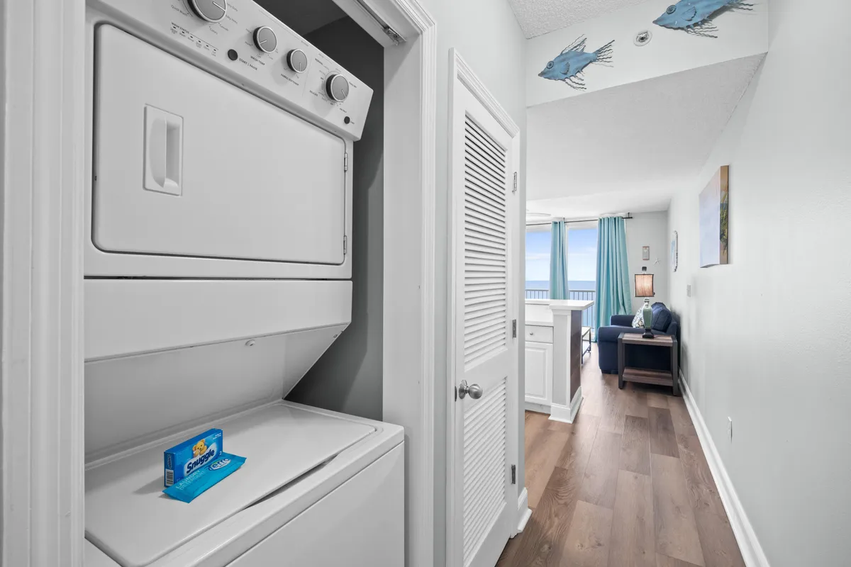 A practical stacked washer and dryer unit is tucked into a hallway that leads to a bright living area with ocean views. Blue curtains frame large windows, and playful fish decorations adorn the ceiling, adding a touch of coastal charm. The layout shows convenient access to laundry within the comfort of the unit.