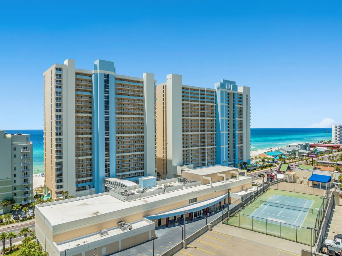 A wide exterior shot of Majestic Beach Resort, showcasing tall high-rise buildings with private balconies overlooking a turquoise ocean. The image prominently features a tennis court in the foreground, multiple resort pools, and recreational areas, all set against a backdrop of white sand and clear skies.