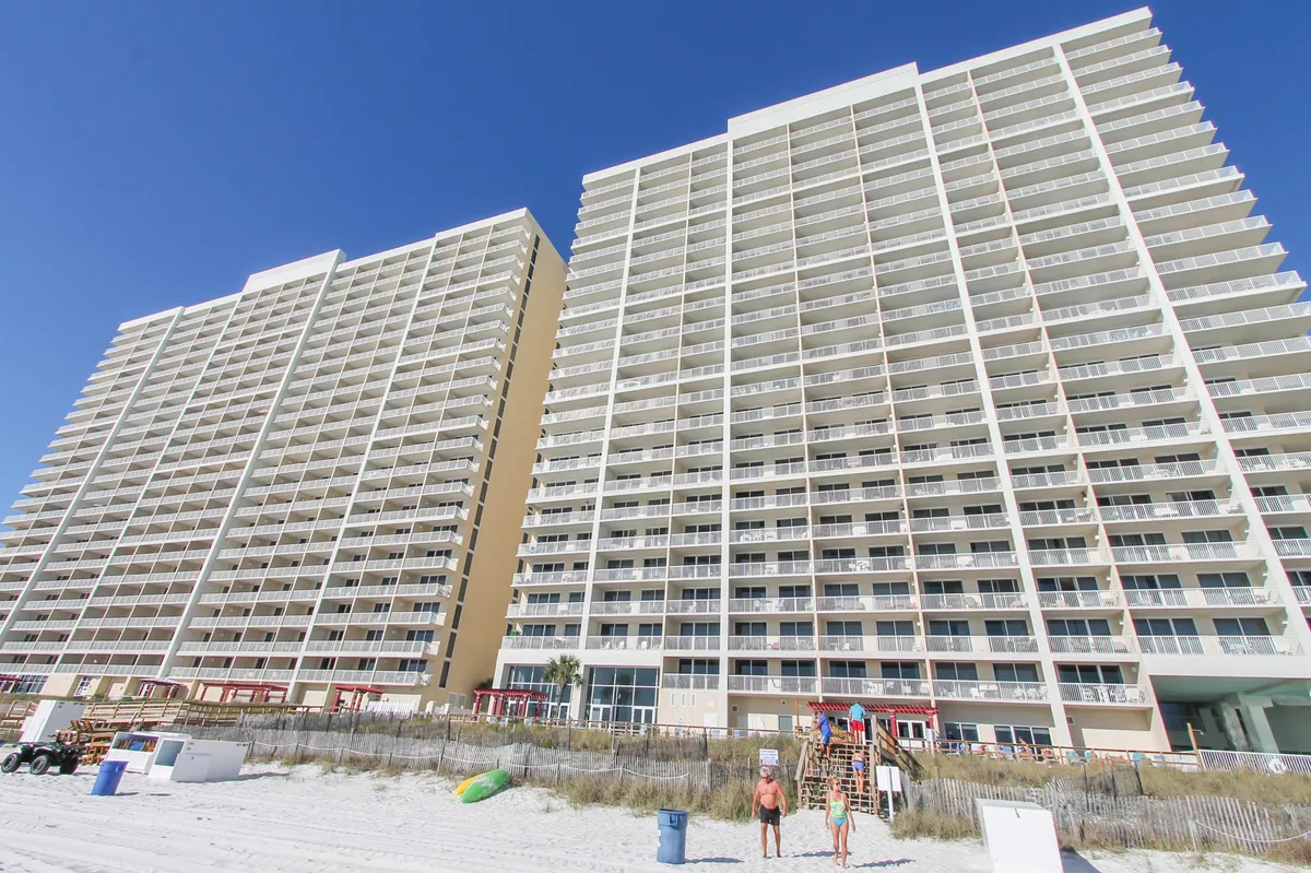 This image showcases tall beachfront condominium buildings with multiple balconies overlooking a sandy beach. There is a small beach access stairway with people visible, and the bright, clear blue sky overhead enhances the relaxed vacation vibe. The scene captures the blend of privacy, ocean proximity, and coastal resort living.