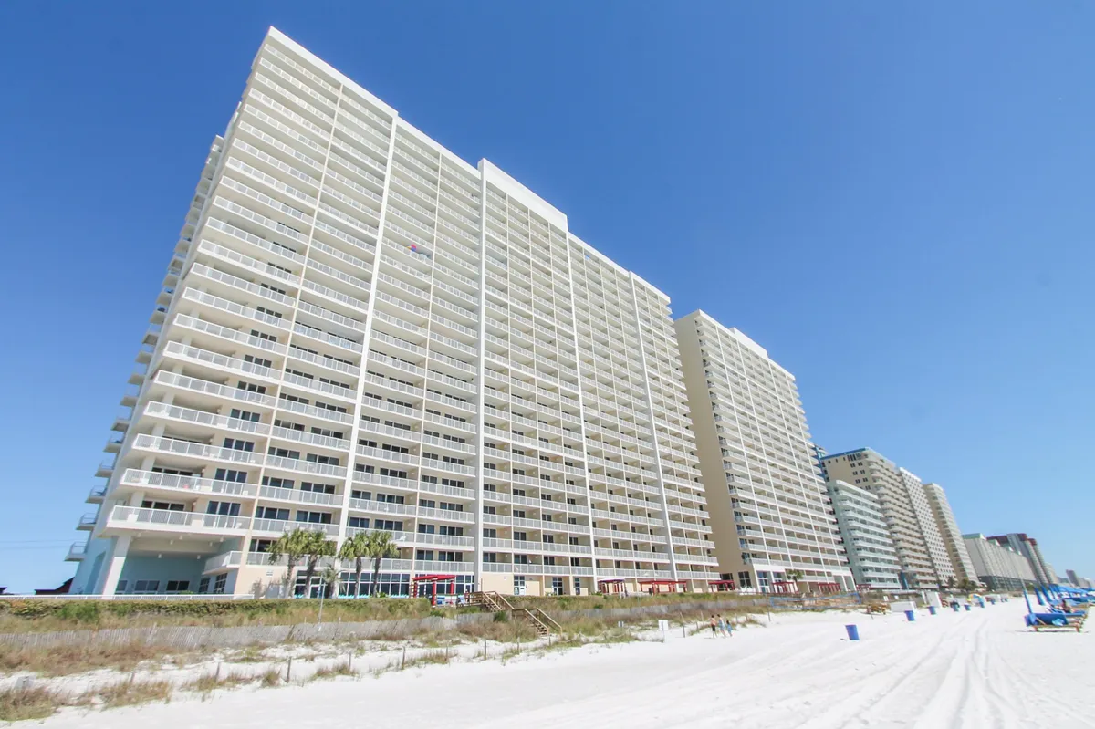 A striking view of the Majestic Beach Resort high-rise, showcasing its numerous private balconies overlooking a pristine white sandy beach with the clear blue sky above. The image hints at the resort’s extensive amenities, with guests able to access pools, tennis courts, and the ocean just steps away.