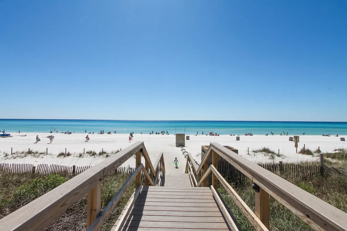A wooden boardwalk leads directly to a white sandy beach with turquoise ocean water. People are seen strolling and enjoying the relaxed beach atmosphere, embodying the leisurely charm of a romantic stroll by the sea.