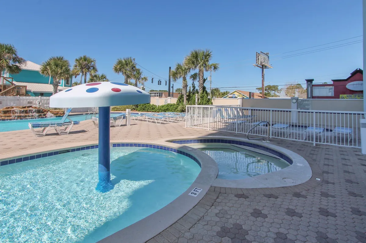 A lively pool area featuring a children’s splash pool with a whimsical mushroom water feature, adjacent to a bubbling hot tub. Lounge chairs and palm trees create a setting for both play and relaxation, with space for families to gather.