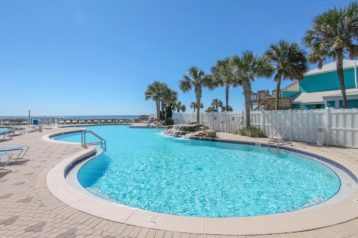 A large, resort-style outdoor swimming pool with a cascading waterfall feature is surrounded by palm trees and lined with sun loungers. The clear blue sky and a distant glimpse of the ocean enhance the vacation ambiance.