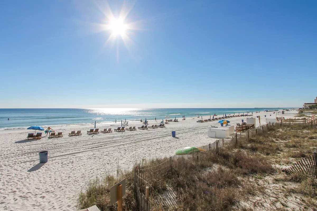A wide sandy beach stretches along the coastline, dotted with lounge chairs and umbrellas. The sparkling ocean water reflects the bright sun, while grass and fencing along the dunes mark the transition from resort to beach. The scene is lively but relaxed, with people enjoying sunbathing, swimming, and the open expanse of the shore.