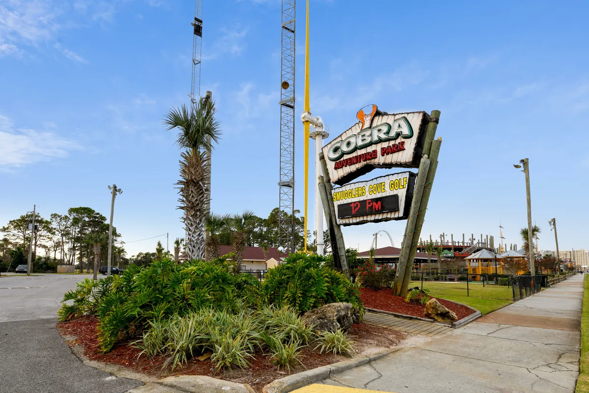 The Cobra Adventure Park sign marks the entrance to a lushly landscaped miniature golf course, with palm trees, vibrant greenery, and adventure rides visible in the background. The scene is playful yet inviting, suggesting fun shared experiences nearby.