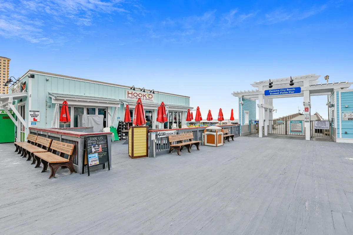 A lively exterior of Hook'd Pier Bar & Grill, with red umbrellas shading outdoor seating and a welcoming boardwalk leading to the entrance archway of the famous Russell Fields Pier. People can be seen enjoying the vibrant, social coastal atmosphere.