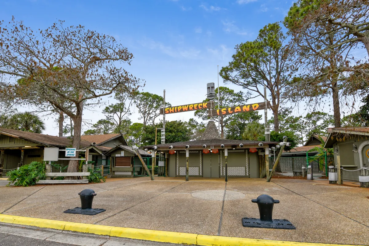 The entrance to Shipwreck Island Waterpark is marked by a rustic wooden sign and surrounding tall trees. Families and groups gather in front, capturing the anticipation and excitement of entering a major local attraction. The open paved area and natural surroundings evoke a sense of adventure and family-friendly fun.